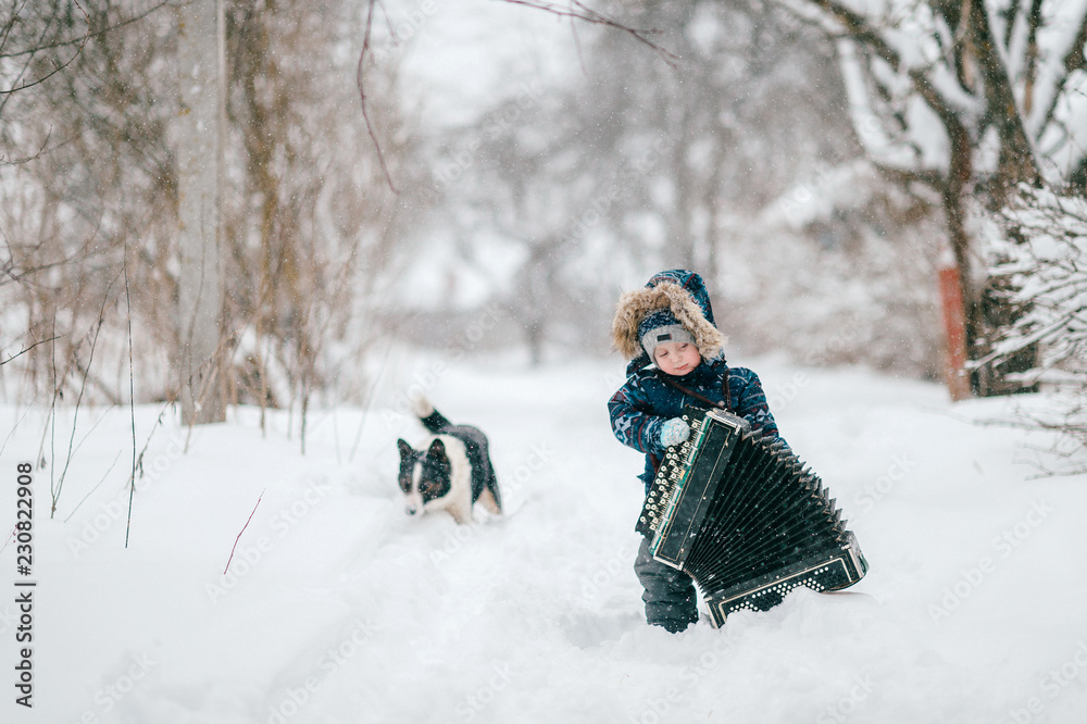 Young happy musician. Comic boy holding big heavy accordion. Lovely funny male kid carrying musician instrument on snowy winter road outdoor. Country lifestyle. Ridiculous baby cold nature portrait.