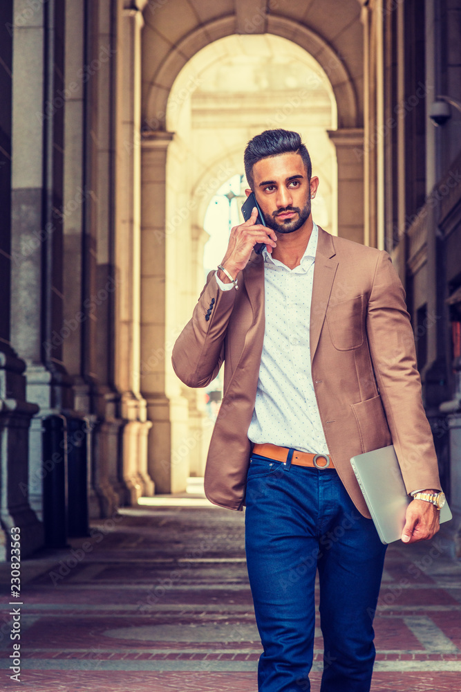 Young East Indian American Businessman traveling, working in New York City,  wearing brown blazer, blue pants, holding laptop computer, talking on cell  phone, walking on vintage narrow street.. Stock Photo | Adobe