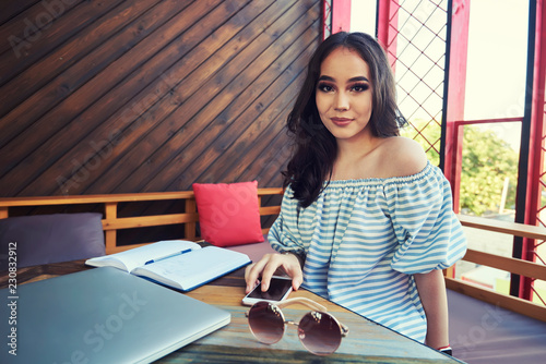 Beautiful cute asian businesswoman in the cafe using mobile phone and drinking coffee. Atractive asin female smiling for someone while is sitting with smarthone and notepad in restaurant photo