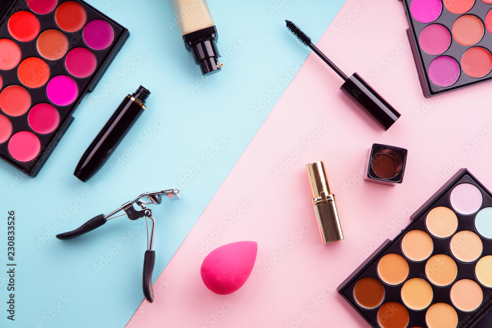 Top view picture of makeup products: lipstick and concealer palettes, foundation, mascara and eyelash curler on pink and blue background. Make-up artist kit concept Top view picture of makeup products: lipstick and concealer palettes, foundation, mascara and eyelash curler on pink and blue background. Make-up artist kit concept