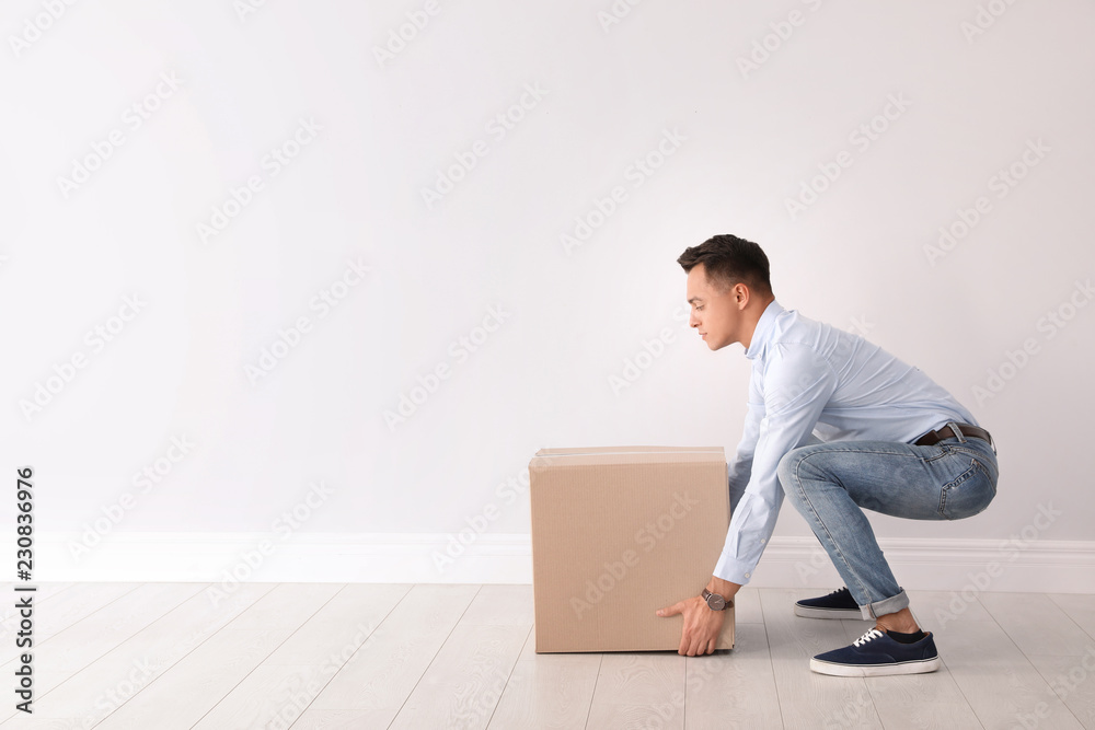 Full length portrait of young man lifting heavy cardboard box near white wall. Posture concept - obrazy, fototapety, plakaty 