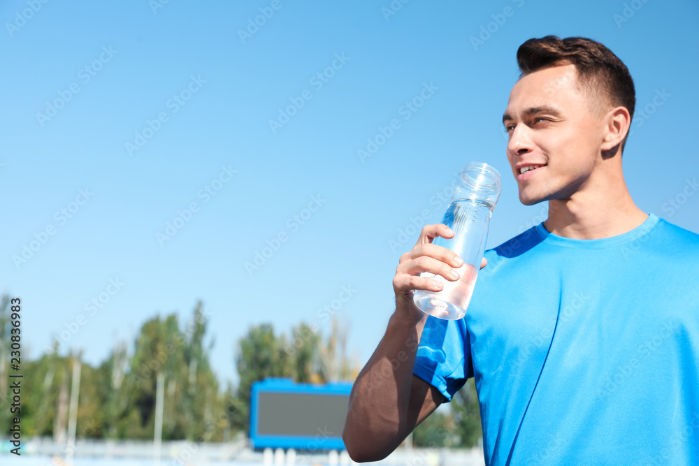 Young sporty man drinking water from bottle at stadium on sunny day. Space for text Young sporty man drinking water from bottle at stadium on sunny day. Space for text