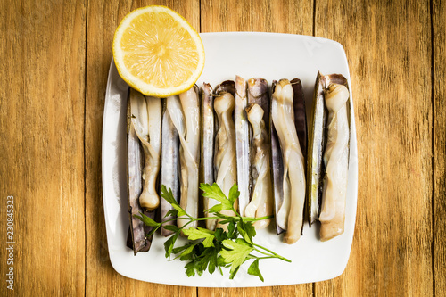 grilled penknives with parsley and half a lemon on a white plate on a wooden table
