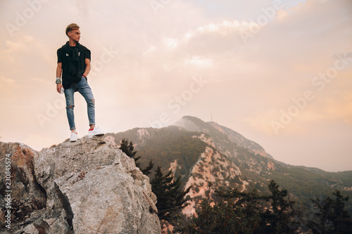 Young man standing on top of cliff in summer mountains at sunset and enjoying view of nature