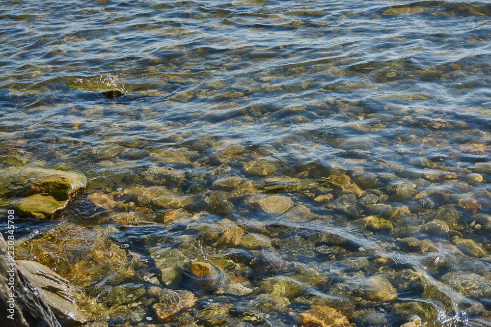 Transparent sea water washes coastal stones overgrown with algae. Stone bottom can be seen through the clear sea surface of water.