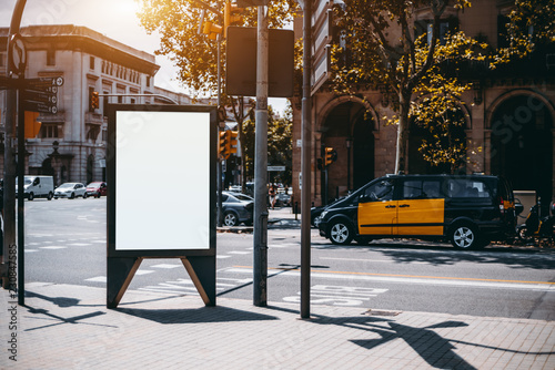 Outdoor empty informational board placeholder with a road and taxi car behind; white blank city billboard mockup; vertical blank advertising banner template on the sidewalk with the crossroad behind