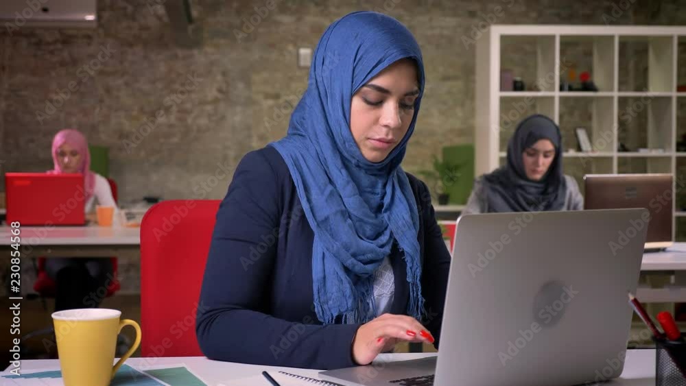 Cute arabic woman in blue hijab is sitting at desktop with colleagues behind and looking cheerful at camera, brick noders office illustration Cute arabic woman in blue hijab is sitting at desktop with colleagues behind and looking cheerful at camera, brick noders office illustration