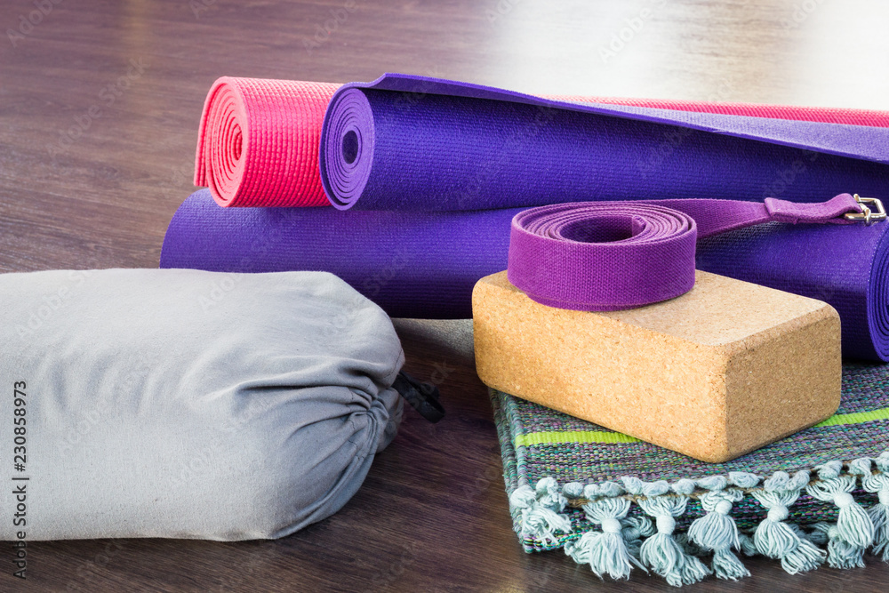 Variety of yoga props on wooden floor in studio. Pink and purple rolled mats, cork brick, belt, grey bolster and folded cotton mat - obrazy, fototapety, plakaty 