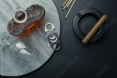 top view of a brandy decanter and a snifter glass with a stainless steel cigar cutter on the marble tray,  matches and a Cuban cigar on the black ceramic ashtray