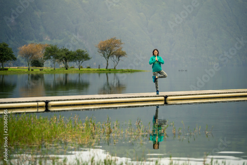 Portrait of young woman in green sweatshirt standing on the edge of pier and meditating. There is volcanic lake on the background. Peaceful atmosphere. Azores islands, Portugal
