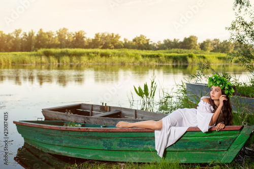 The nymph with long dark hair in a white vintage dress sitting in a boat in the middle of the river. photo