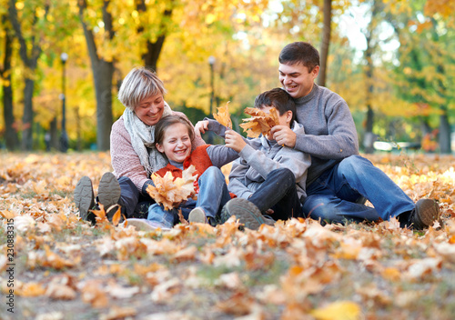 Happy family sit in autumn city park on fallen leaves. Children and parents posing, smiling, playing and having fun. Bright yellow trees.
