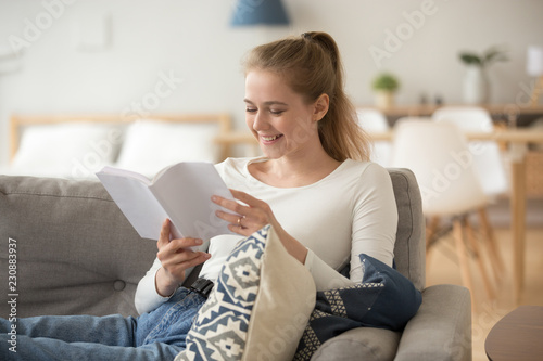 Beautiful cheerful woman spends free time on weekend alone sitting on couch in living room holding a book at modern comfortable home. Young girl laughing enjoying reading love story novel or chicklit photo