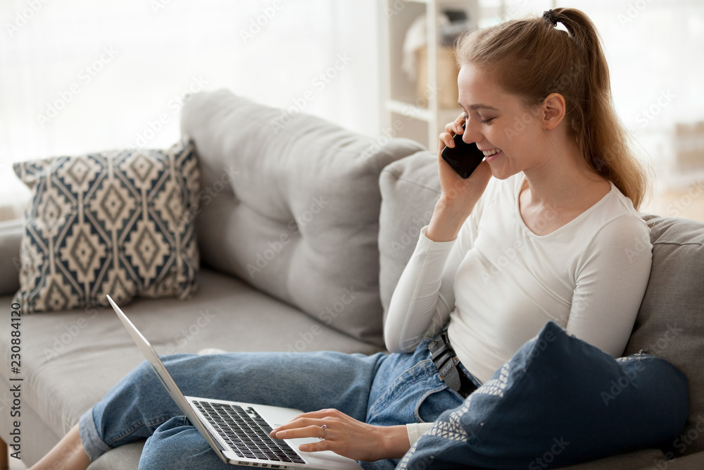 Happy woman sitting on couch in living room at home alone typing chatting using computer and talking by phone. Positive female makes an order via internet, cheerful girl has conversation with friend Happy woman sitting on couch in living room at home alone typing chatting using computer and talking by phone. Positive female makes an order via internet, cheerful girl has conversation with friend