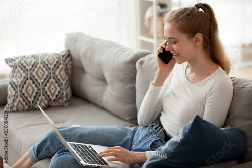 Happy woman sitting on couch in living room at home alone typing chatting using computer and talking by phone. Positive female makes an order via internet, cheerful girl has conversation with friend