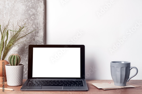 Hipster desk with a mockup laptop; a cup of coffee, a cactus in a gray pot; a vase with green grass and a stone on the wall with copy space