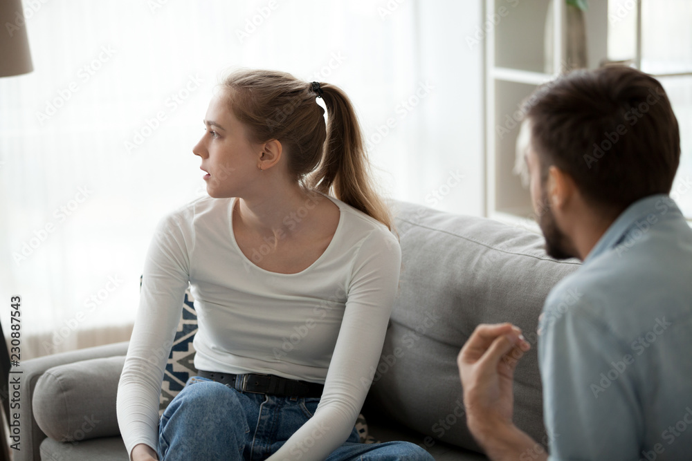 Unhappy couple sitting on couch in living room at home sorting out their relationships quarrelling. Guy arguing emotionally talking frustrated girl looking away. Break up problems in relations concept