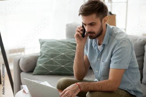 Man sitting on couch reading message email online looking at laptop talking by mobile phone. Male working at home has serious conversation with client. Businessman solve problems and matters by phone