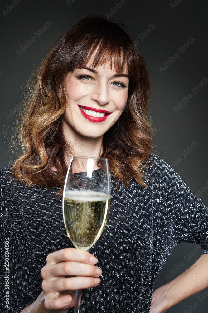 Woman wearing night party dress with a glass of champagne on dark background. Lady with long curly hair and perfect teeth celebrating and smiling. Woman wearing night party dress with a glass of champagne on dark background. Lady with long curly hair and perfect teeth celebrating and smiling.