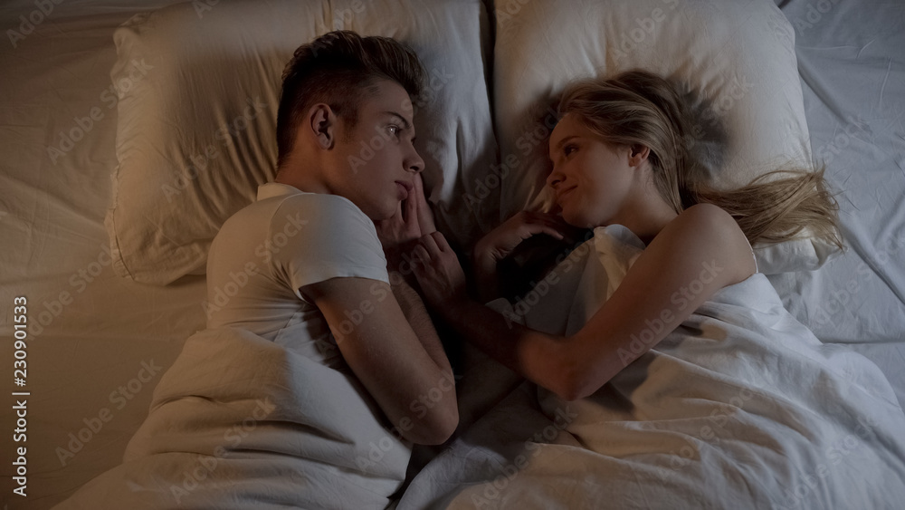 Smiling newlyweds looking each other in bed, holding hands, relax on soft linens Smiling newlyweds looking each other in bed, holding hands, relax on soft linens
