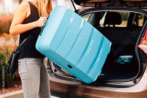 Young woman loading two blue plastic suitcases to car trunk.