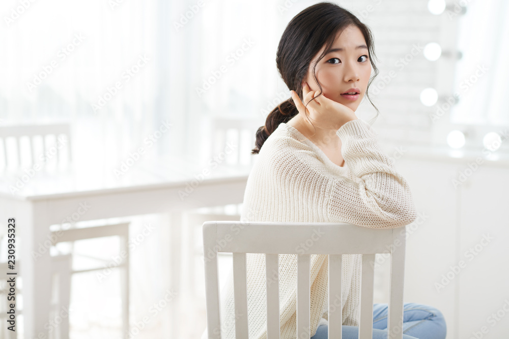 Beautiful young Korean woman sitting in white room and turning back to camera Beautiful young Korean woman sitting in white room and turning back to camera