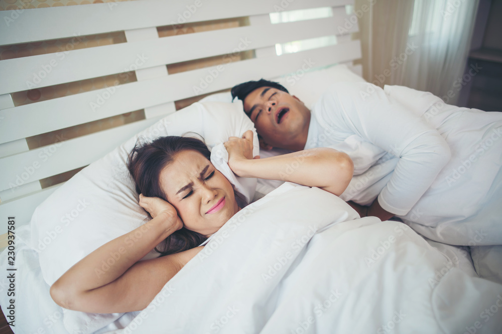 Fototapeta premium Portrait of woman blocking ears with man sleeping snoring on bed