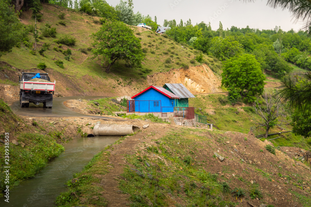 A blue colour hut, water canal, a truck moving upslope on a road A blue colour hut, water canal, a truck moving upslope on a road