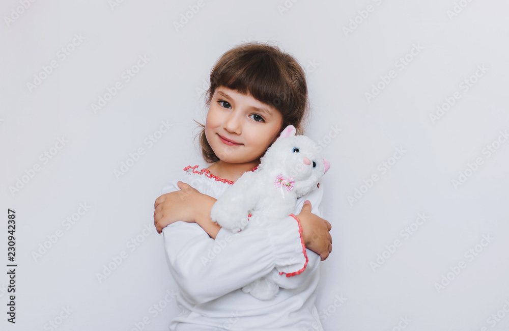 Happy little girl hugging her favorite white kitten soft toy on a white back. Favorite toys of children. Orphanage. Copy space. Happy little girl hugging her favorite white kitten soft toy on a white back. Favorite toys of children. Orphanage. Copy space.