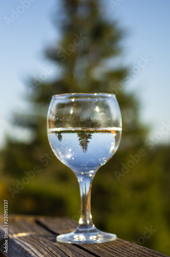 A glass with a water on the balcony overlooking the mountains. Mirror display in a glass. Display a conifer tree in a glass