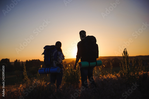 Silhouettes of two hikers with backpacks walking at sunset. Trekking and enjoying the sunset view