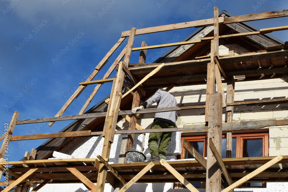 Worker working on wooden scaffolding and installing rigid  polystyrene styrofoam plates on residential building facade. House insulation improvement process nd DIY concept.