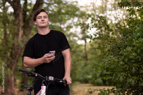 Attractive young caucasian man holds mobile phone and bicycle. White earphones, earring, dark casual wear, dark hair and eyes. Stands at the abandoned park. Outdoors, close up, copy space.