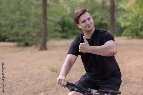Handsome young caucasian man thumbs up biking at the park. White earphones, dark casual wear. Outdoors, abandoned park. Close up, copy space. Smile, emotions and expression. Diversity people.