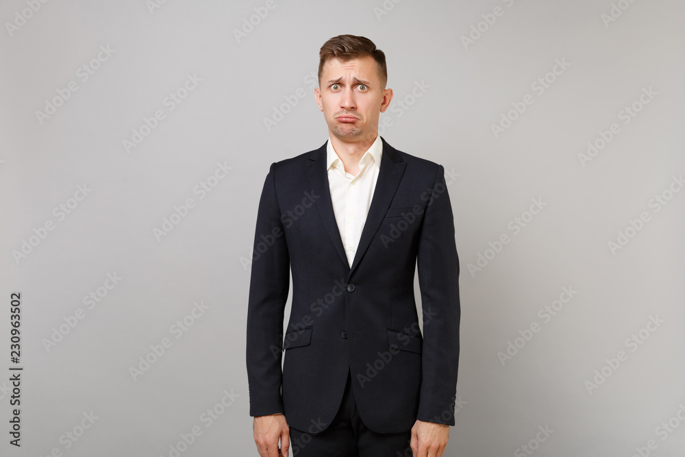 Fototapeta premium Portrait of offended inflated young business man in classic black suit, white shirt standing isolated on grey wall background in studio. Achievement career wealth business concept. Mock up copy space.