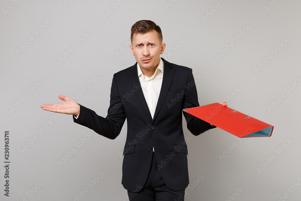 Puzzled young business man in classic black suit, shirt spreading hands with red folder for papers document isolated on grey background. Achievement career wealth business concept. Mock up copy space. Puzzled young business man in classic black suit, shirt spreading hands with red folder for papers document isolated on grey background. Achievement career wealth business concept. Mock up copy space.