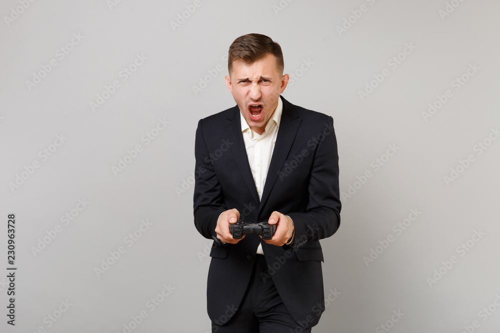 Portrait of emotional young business man in classic black suit, shirt playing video game with joystick isolated on grey wall background. Achievement career wealth business concept. Mock up copy space. Portrait of emotional young business man in classic black suit, shirt playing video game with joystick isolated on grey wall background. Achievement career wealth business concept. Mock up copy space.