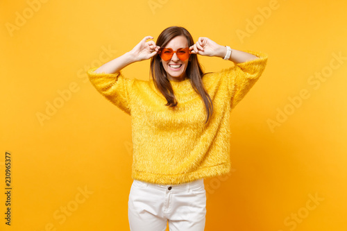 Portrait of laughing happy young woman in fur sweater, white pants holding heart orange eyeglasses isolated on bright yellow background. People sincere emotions, lifestyle concept. Advertising area.