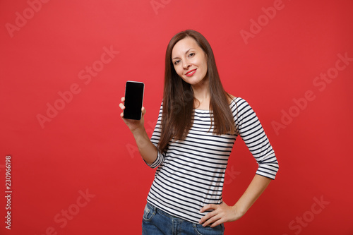 Portrait of stunning young woman in striped clothes holding mobile phone with blank black empty screen isolated on bright red background. People sincere emotions lifestyle concept. Mock up copy space.