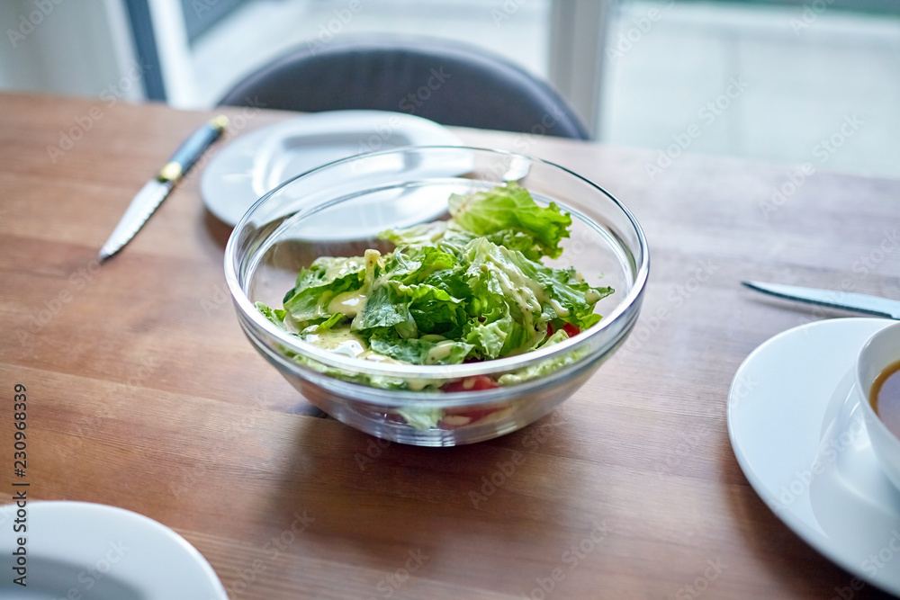 Fresh Vegetable Dish. Vegetables in a transparent bowl on a wooden dinner table. Ingredients: slices of red tomato and green salad leaves sprinkled with olive (or sunflower) oil and sauce Fresh Vegetable Dish. Vegetables in a transparent bowl on a wooden dinner table. Ingredients: slices of red tomato and green salad leaves sprinkled with olive (or sunflower) oil and sauce