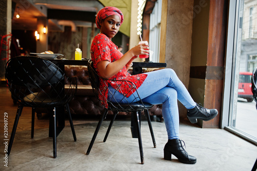 Stylish african woman in red shirt and hat posed indoor cafe and drinking strawberry lemonade with phone at hand.