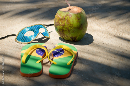 Brazil flag flip flops sit next to a carnival mask with a green coconut under the shadows of palm fronds on a Brazilian beach