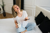 Happy young beautiful woman drinking coffee in the morning, sitting on the bed in her pajamas and laughing out loud because she read something funny on the phone.