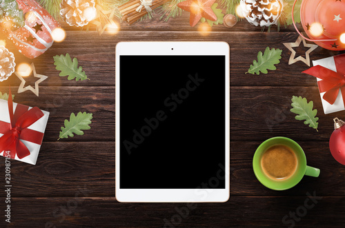 Digital tablet with blank screen surrounded with Christmas decorations and coffee on dark wooden desk for app presentation, top view