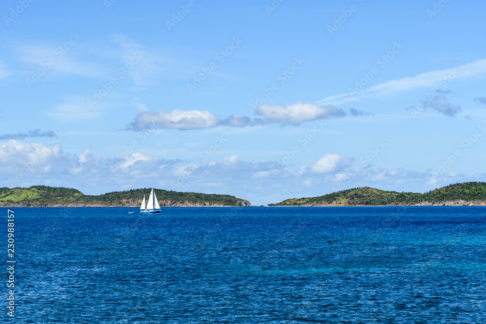 Yacht with white sails on the blue sea against the green shore and clouds