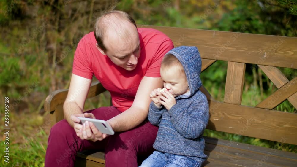 The boy gives the man a bite wound. Son plays Apple with father. A man with a child using a smartphone sitting on a bench. 4k The boy gives the man a bite wound. Son plays Apple with father. A man with a child using a smartphone sitting on a bench. 4k