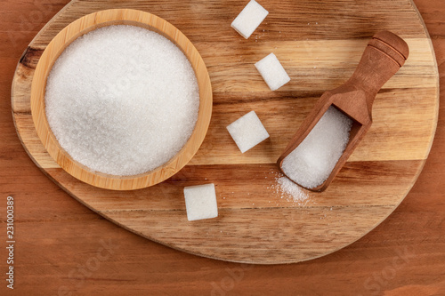 An overhead photo of a bowl and a scoop of white sugar, with sugar cubes, shot from the top on dark rustic wooden backgrounds with copy space