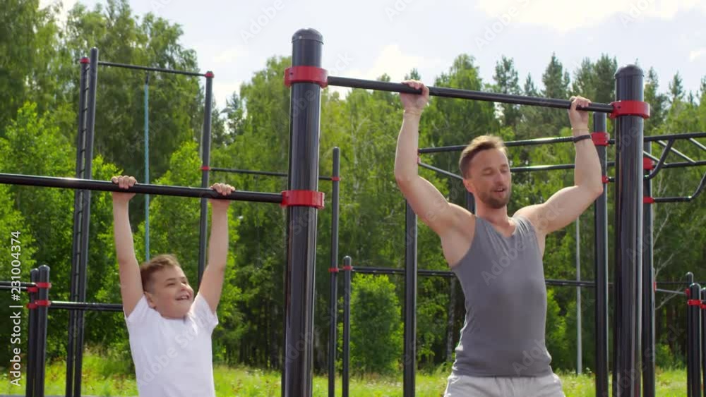 Athletic father and little son doing pull ups on bars while training together at outdoor gym zone in the park on summer day Athletic father and little son doing pull ups on bars while training together at outdoor gym zone in the park on summer day