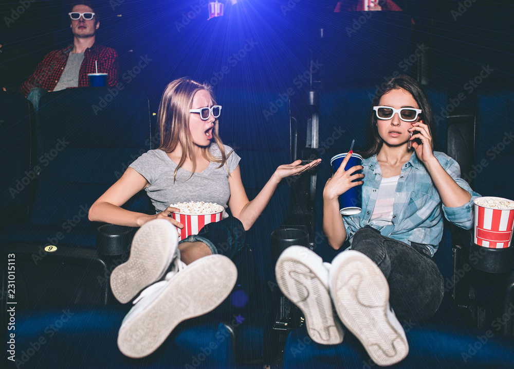 Fototapeta premium Girls are sitting in chairs in cinema hall. Brunette is talking on the phone while her friend is making remark to her. Girl on th left is upset and irritated.