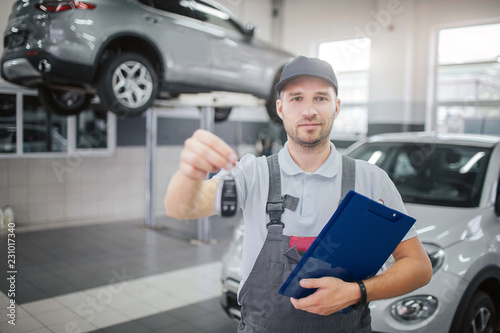 Nice and confident man stands and looks on camera. He holds car keys and plastic folder with documents. There are tow cars behind him. © estradaanton